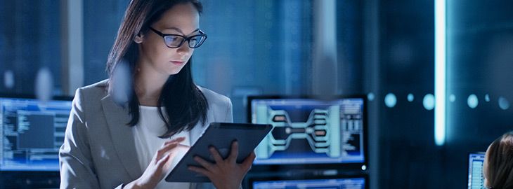 Young Female Government Employee Wearing Glasses Uses Tablet in System Control Center. In the Background Her Coworkers are at Their Workspaces with many Displays Showing Valuable Data.